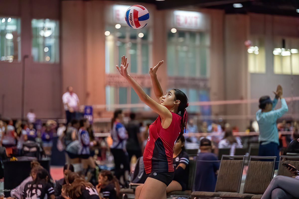A volleyball player in a red jersey serves the ball at an indoor court, with other players and spectators in the background.