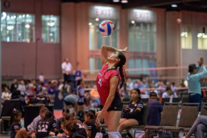 A volleyball player in a red jersey jumps to hit the ball during an indoor game while teammates and spectators watch.