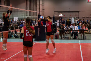 A volleyball player in a red jersey receives the ball during an indoor game. Another player in a red jersey stands nearby, and an opposing player in a black jersey attempts to block at the net.