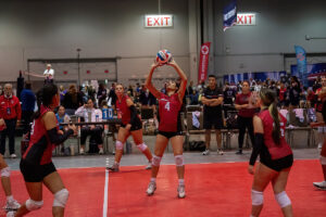 A volleyball game inside a sports hall. A player in a red jersey is setting the ball while teammates and spectators watch. Two exit signs and various banners are visible in the background.