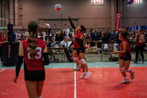 A volleyball player jumps to hit a ball while three teammates and a coach observe during an indoor volleyball match. The background shows the audience and exit signs.