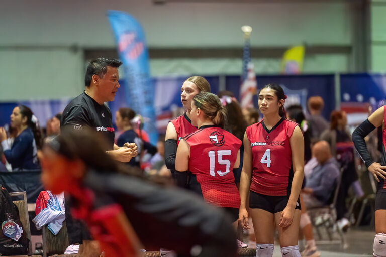 A volleyball coach speaks to four players in red jerseys, identified by numbers 4 and 15, during an indoor tournament. Other players and spectators are visible in the background.