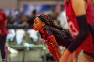 A focused volleyball player bends forward, resting her hands on her knees, while observing the action on the court. She is wearing a black jersey with red accents and a headband.