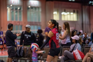 A volleyball player in a red and black jersey prepares to serve, holding the ball in one hand. Spectators in the background are watching and conversing.
