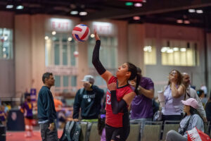 A volleyball player in a red jersey serves the ball during an indoor game as spectators watch from the sidelines.