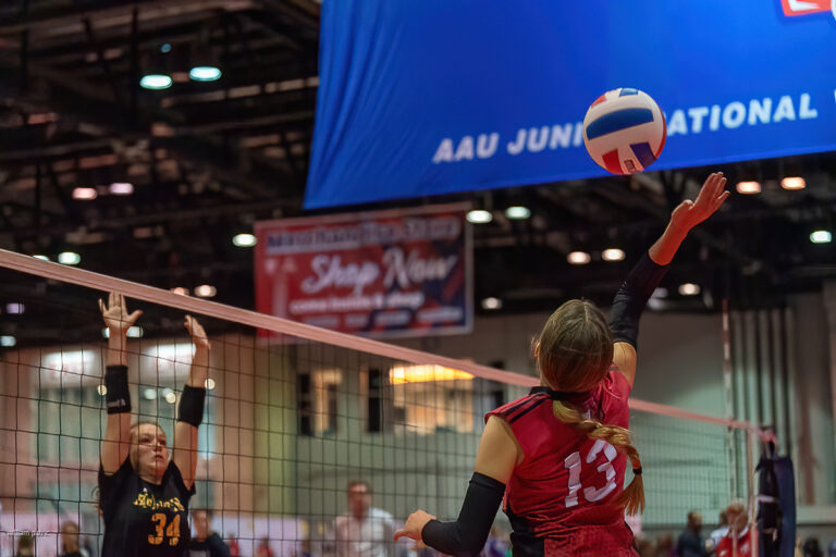 A volleyball player in a red jersey jumps to hit the ball while a player in a black jersey attempts to block at the net during a match at the AAU Junior National Championship.
