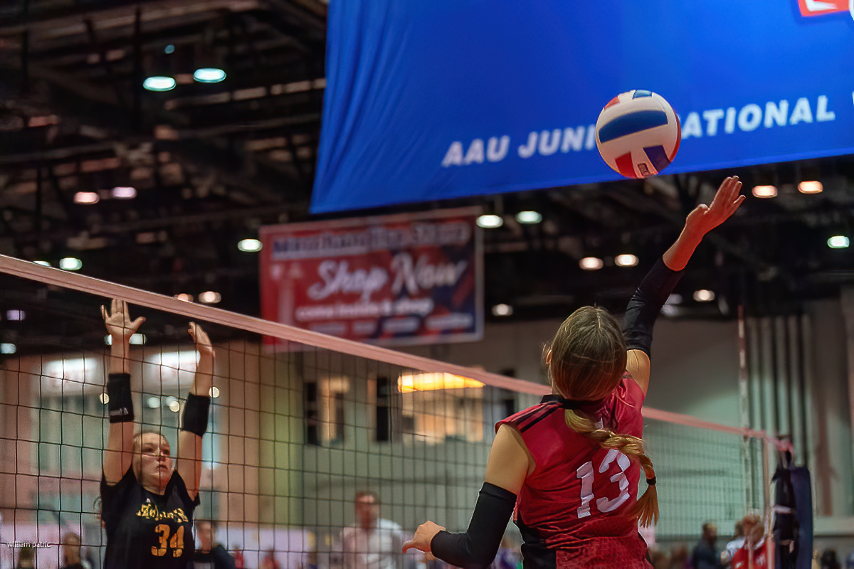 A volleyball player in a red jersey jumps to hit the ball while a player in a black jersey attempts to block at the net during a match at the AAU Junior National Championship.