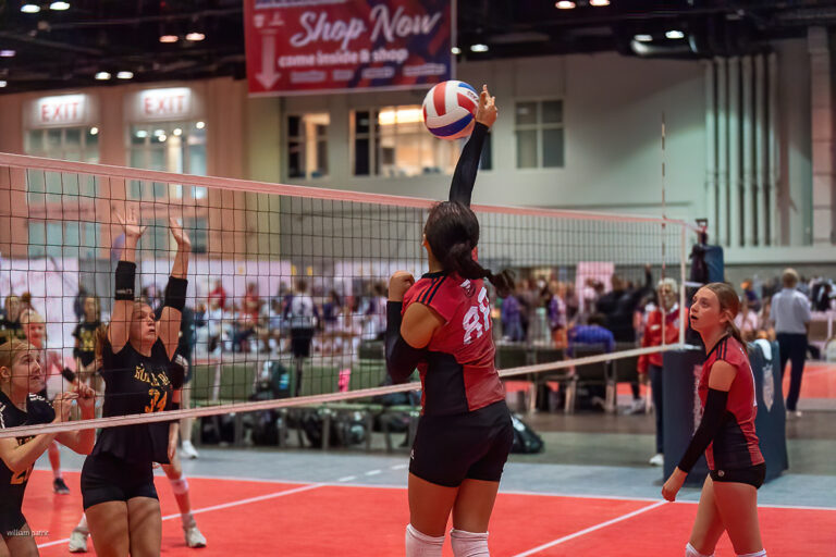 A volleyball player in a red and black uniform jumps to spike the ball over the net while an opposing player in a yellow and black uniform attempts to block.