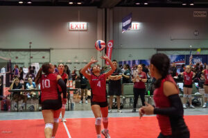 Indoor volleyball game with a player wearing number 4 preparing to set the ball while teammates wearing red jerseys wait in a sports facility with an exit sign in the background.