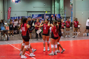 A women's volleyball team celebrates on the court in front of a scoreboard showing 13-12. Several players are clapping and high-fiving while others look on. The venue is an indoor sports facility.