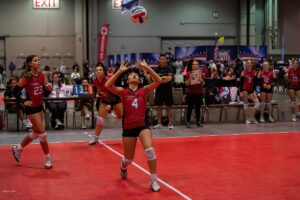 A volleyball player in a red jersey prepares to set the ball during an indoor game. Teammates and officials watch from the sidelines, with a scoreboard displaying 13 to 2 in the background.
