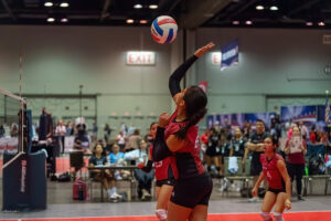 A volleyball player in a red and black uniform is jumping to spike the ball during an indoor game. Teammates and spectators are visible in the background.