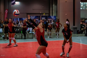 A volleyball player in a red jersey prepares to hit a ball, while a teammate and opponents look on in the background. The game takes place on an indoor court during a match.