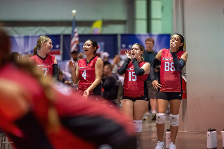 Four players on a volleyball team in red uniforms are standing on the sidelines, cheering and smiling.
