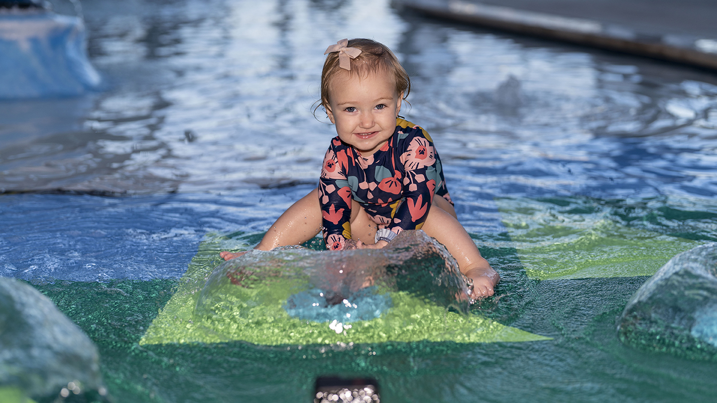 A toddler wearing a floral swimsuit and a hair bow sits and smiles on a water play area with shallow water, capturing the perfect moment for event photography.