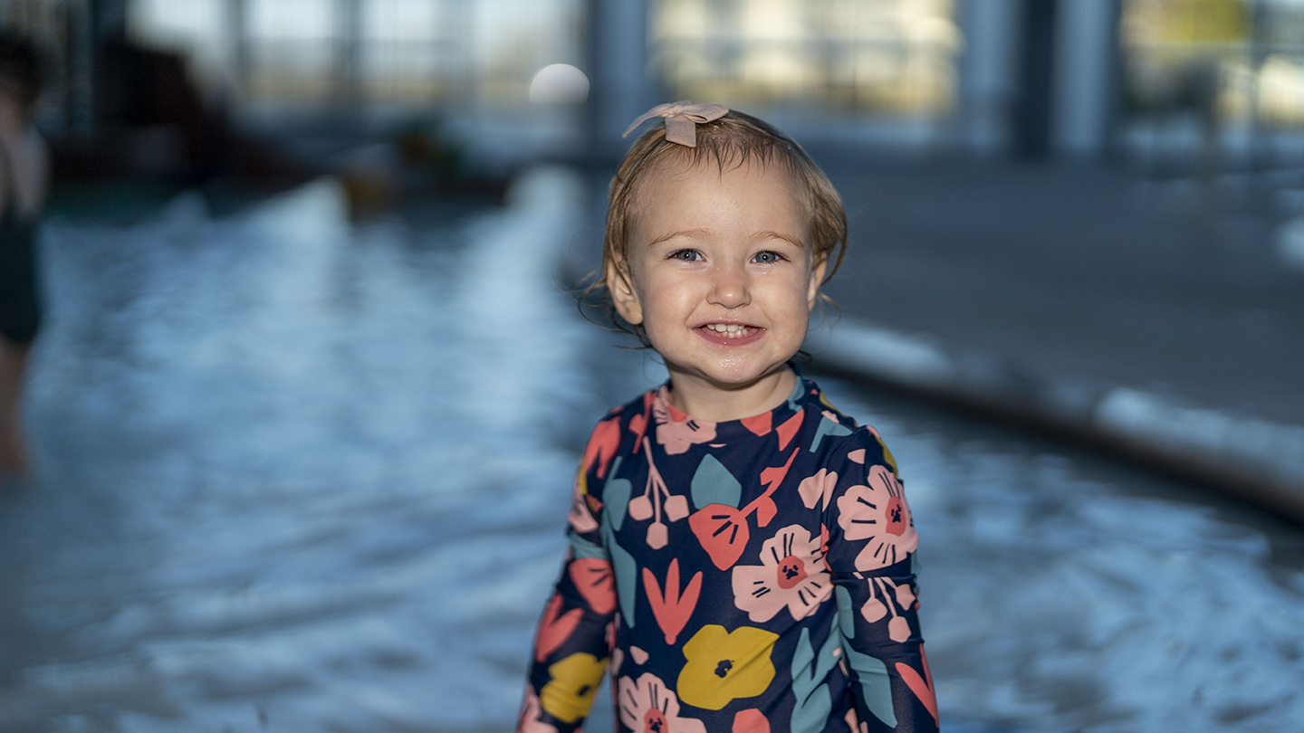 A young child with a bow in her hair smiles while standing in front of a pool, captured beautifully through event photography. She is wearing a floral-patterned dress.