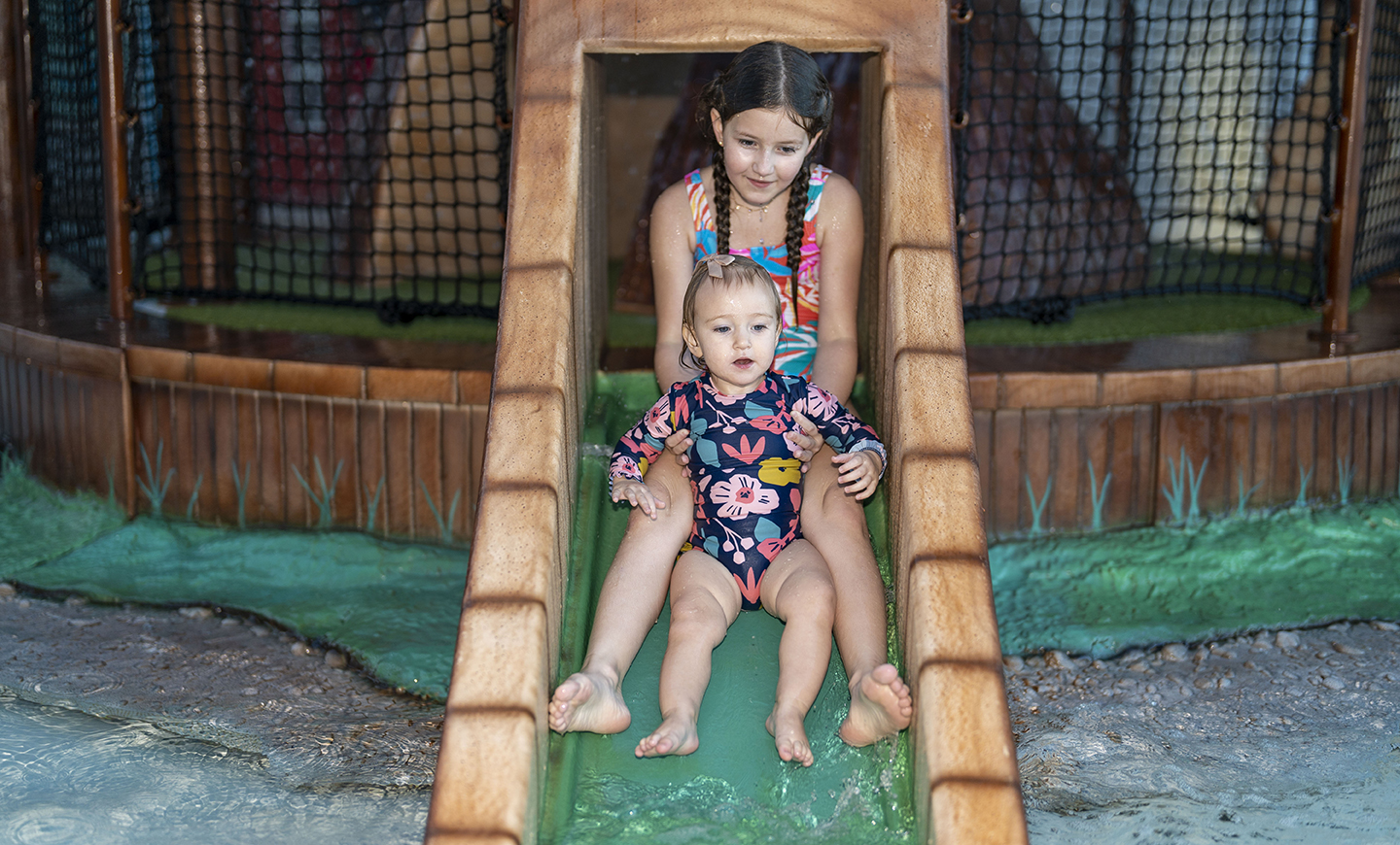 Two young children in swimsuits gleefully slide down a playground slide with a protective mesh enclosure, perfectly capturing the joy of summer fun and the essence of event photography.