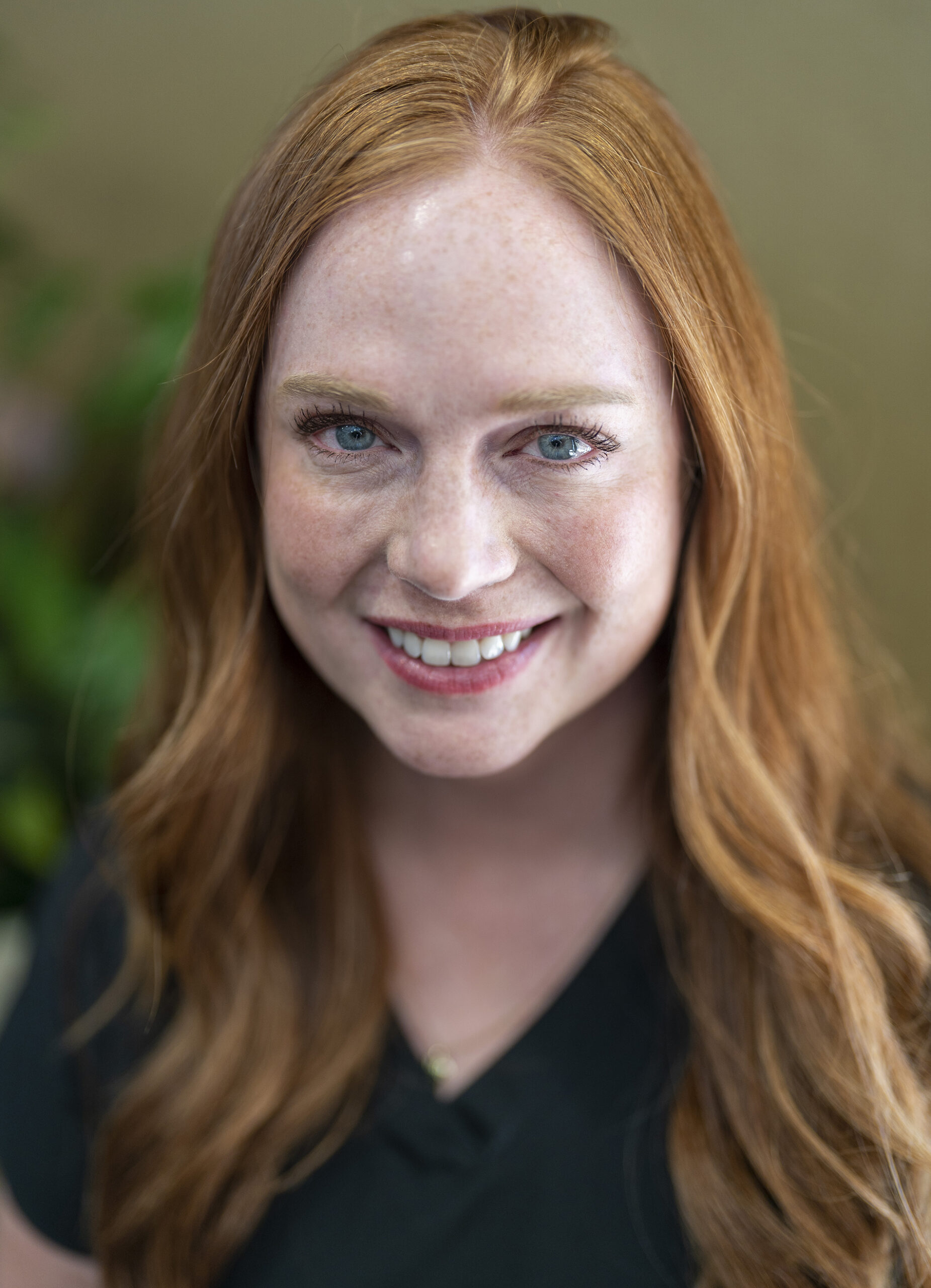 A woman with long red hair and freckles smiles directly at the camera. She is wearing a dark green top.