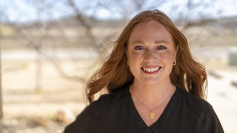 Red-haired woman smiling outdoors, wearing a black top and gold necklace. Trees and a blurred background are visible behind her.