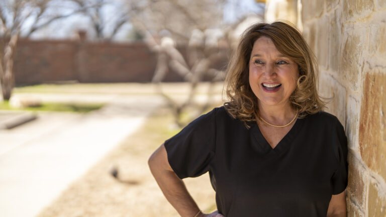 A woman in a black blouse stands outdoors by a stone wall, smiling and looking to the side. The background shows a yard with bare trees and a brick wall.