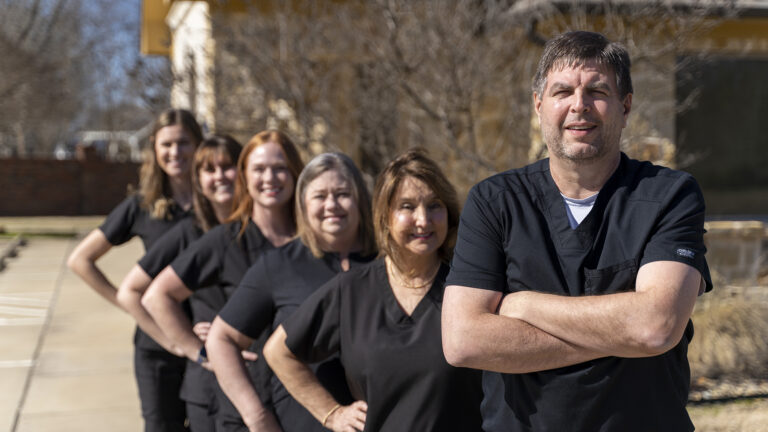 A group of six people wearing black uniforms stand in an outdoor setting. The person in the foreground has their arms crossed, while the others are standing in a line behind.