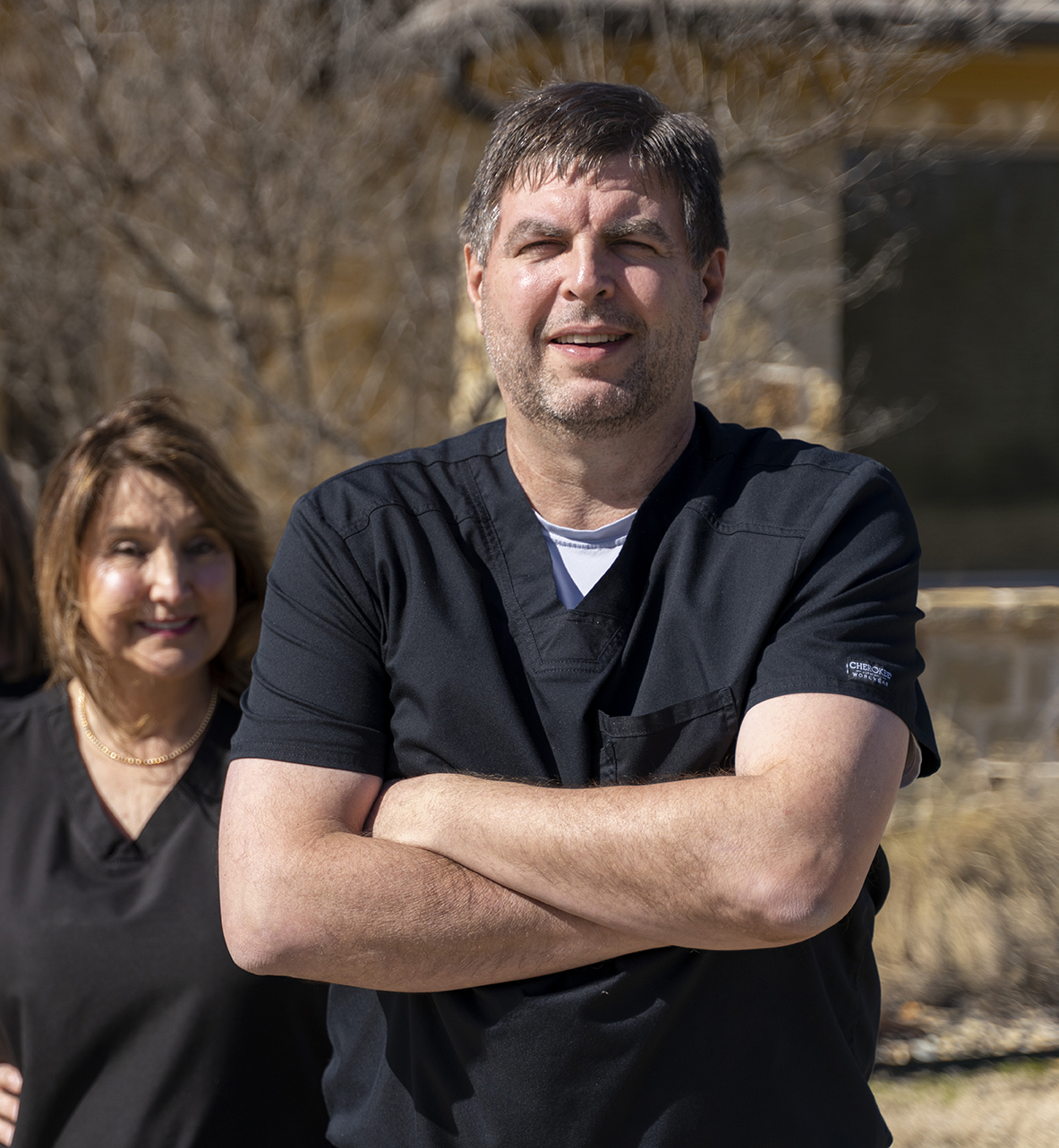 A man stands outdoors with his arms crossed, wearing black medical scrubs. A woman in similar attire stands slightly behind him. They appear to be in a sunny, wooded area.