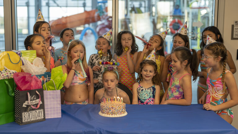 A girl in front of a birthday cake with lit candles is surrounded by children wearing party hats and blowing party horns, standing around a table covered with a blue cloth and gift bags – a perfect scene for event photography.