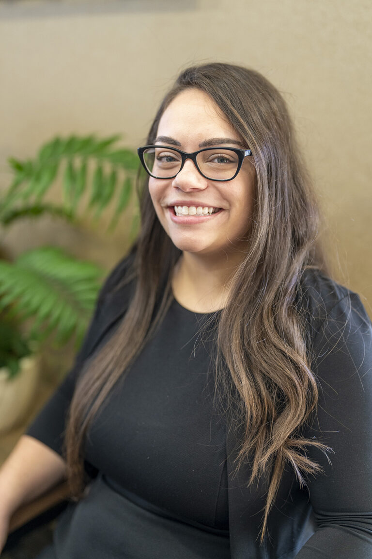 A woman with long brown hair and glasses, wearing a black top, smiles while seated indoors. A green plant is visible in the background.