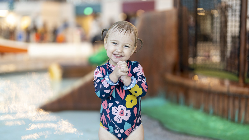A young child with pigtails smiles while standing in a water play area, wearing a long-sleeved floral swimsuit. Perfect for event photography, this candid moment captures the joy and innocence of childhood.