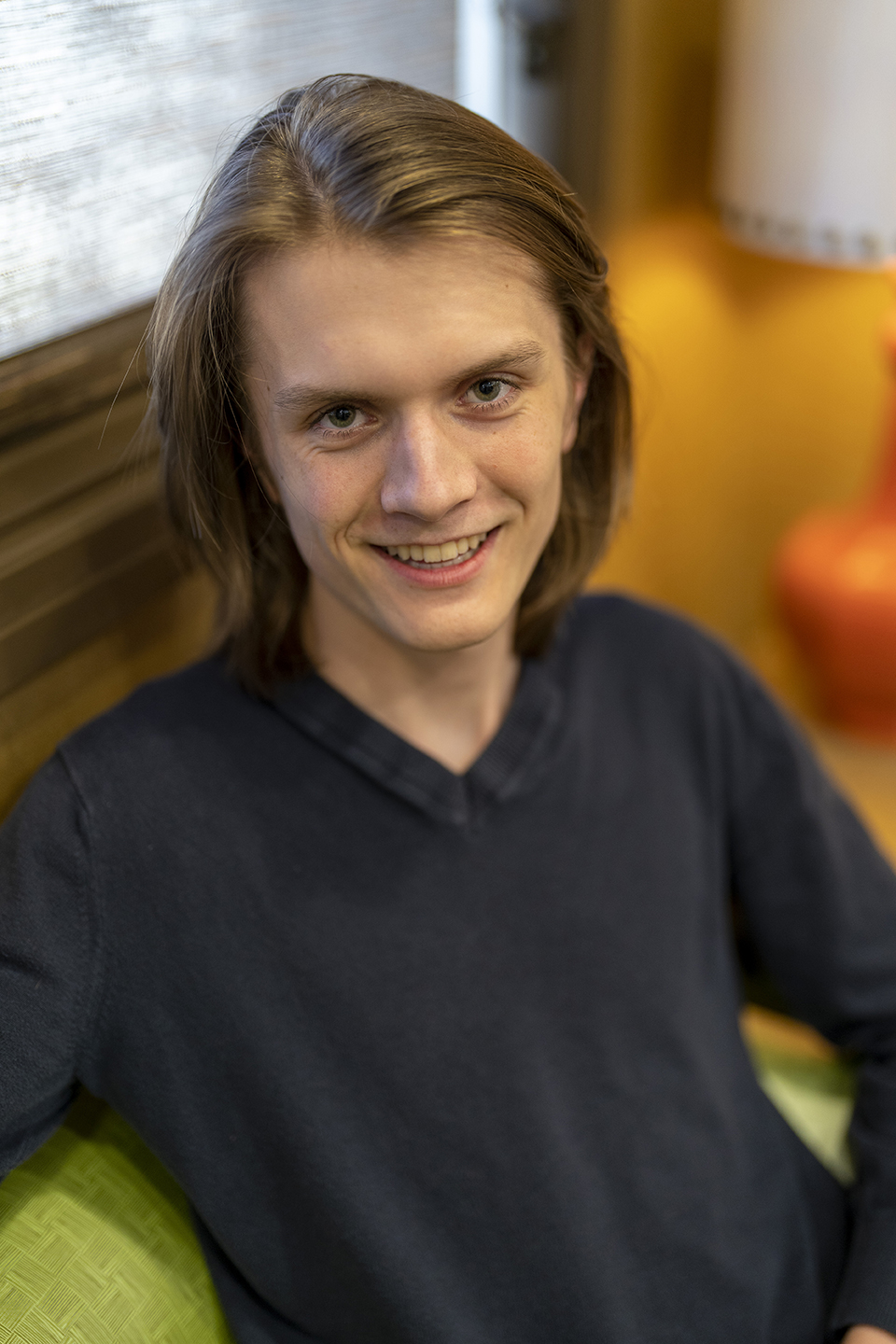 A person with shoulder-length hair wearing a dark long-sleeve shirt sits on a green chair indoors, smiling at the camera.