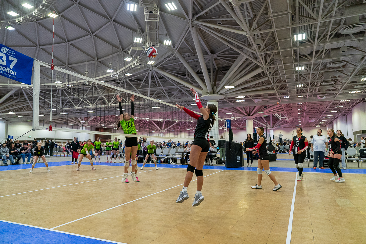 Indoor volleyball match with players jumping and reaching for the ball under a high ceiling, while a crowd watches from the background.