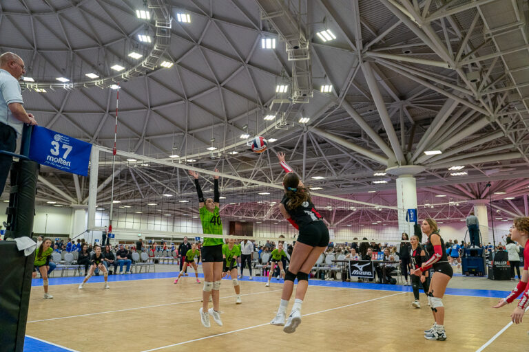 Indoor volleyball game with players in mid-air action near the net. Referee watches from a stand, while a crowd observes from the sidelines in a sports arena.