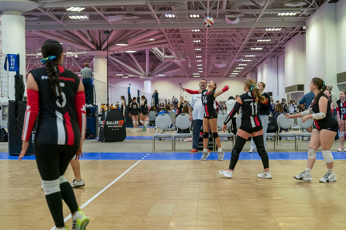 Volleyball players on an indoor court during a match, with one player preparing to serve while teammates and opponents are positioned and watching.