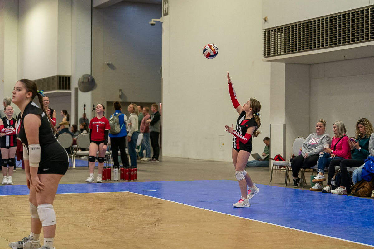 A young volleyball player in a red and black uniform serves the ball indoors, while teammates and spectators watch from the sidelines.