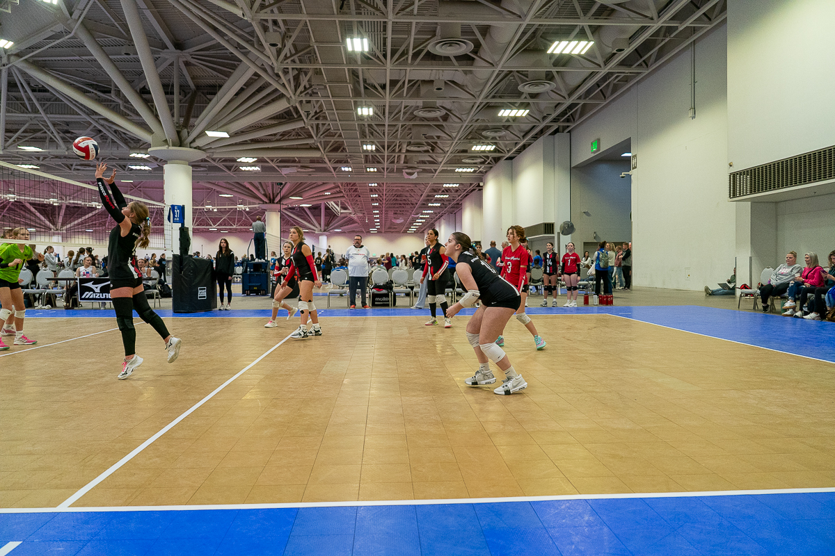 Volleyball players in action on an indoor court, with one player jumping to hit the ball. Spectators watch from the sidelines.