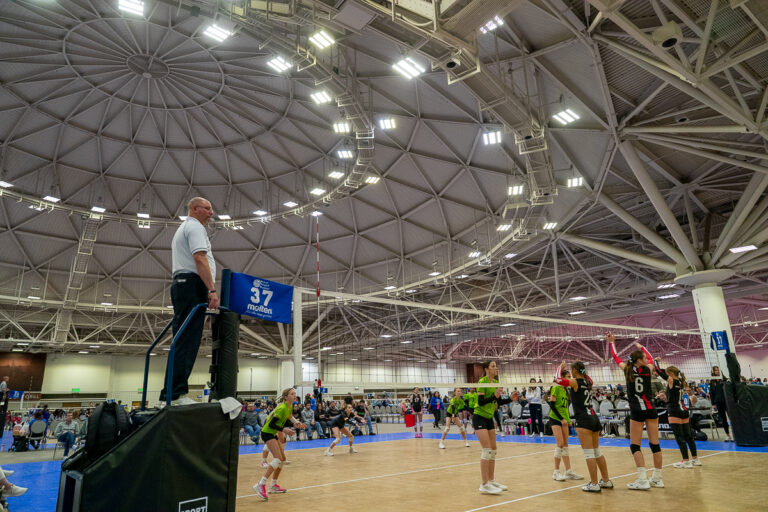Volleyball match in a large indoor stadium with players on the court and a referee standing on a platform. Spectators are seated in the background.