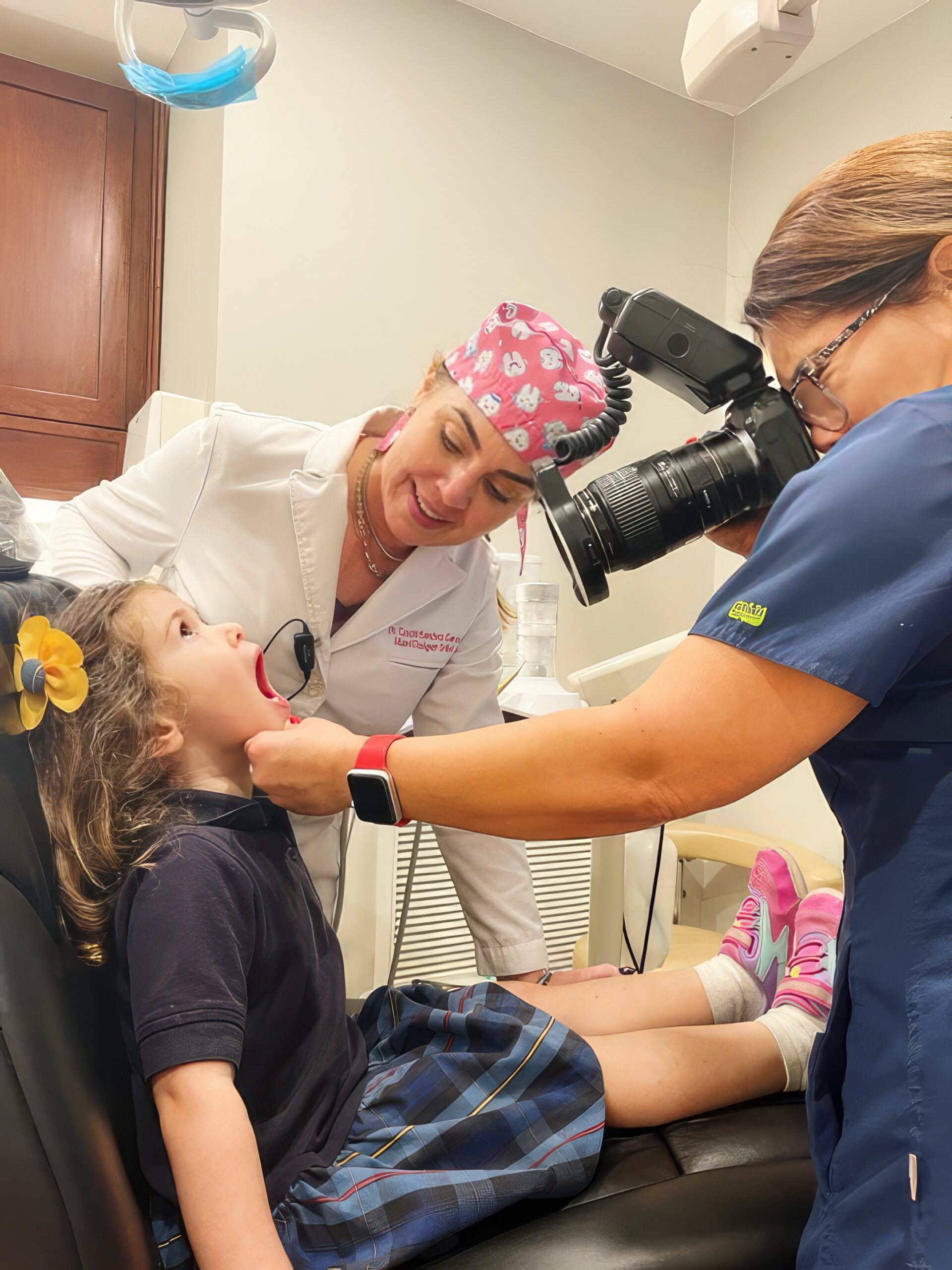 A child sits in a dental chair with an open mouth. A dentist and an assistant, who is capturing a photo, explore the evolving trends in pediatric dentistry through this candid portrait.