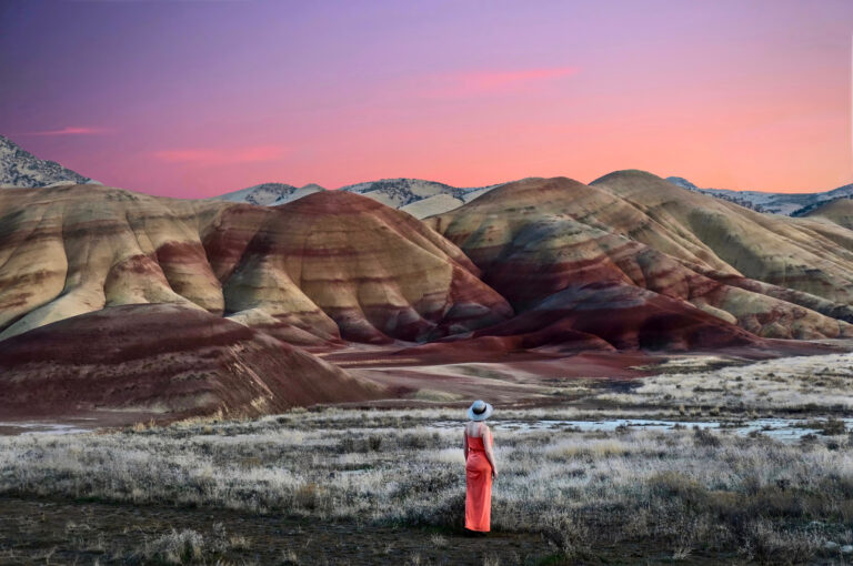 A person in a coral dress and hat stands facing colorful, striped hills at sunset, capturing one of the most stunning photo spots perfect for a spring break adventure, with a vivid gradient sky adding to the breathtaking view.