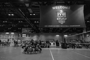 A volleyball team huddles on an indoor court beneath a large banner reading "Welcome AAU Volleyball AAU Junior National Volleyball Championship," captured in striking black and white sports photography.
