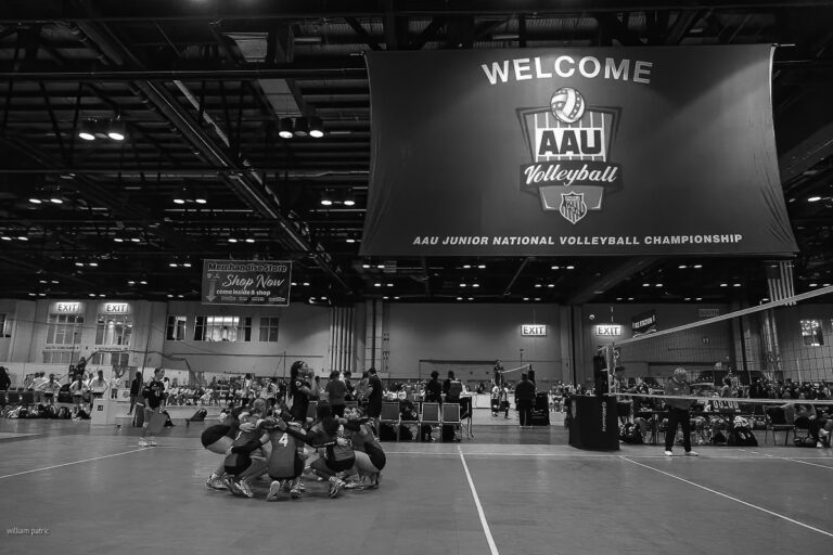 A volleyball team huddles on an indoor court beneath a large banner reading "Welcome AAU Volleyball AAU Junior National Volleyball Championship," captured in striking black and white sports photography.