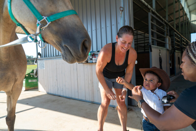 A woman and a child in a cowboy hat interact with another woman beside a horse at a stable. The child appears hesitant while being offered something.