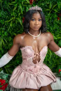 A woman in a pink tiara, pearl necklace, and pink strapless dress sits in front of a green leafy backdrop, looking at the camera.