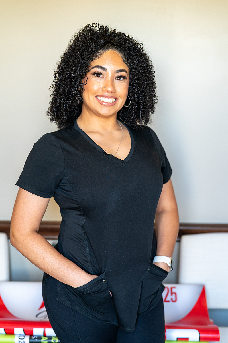 A woman with curly hair, wearing a black scrub top, stands indoors with her hands in her pockets, smiling at the camera.