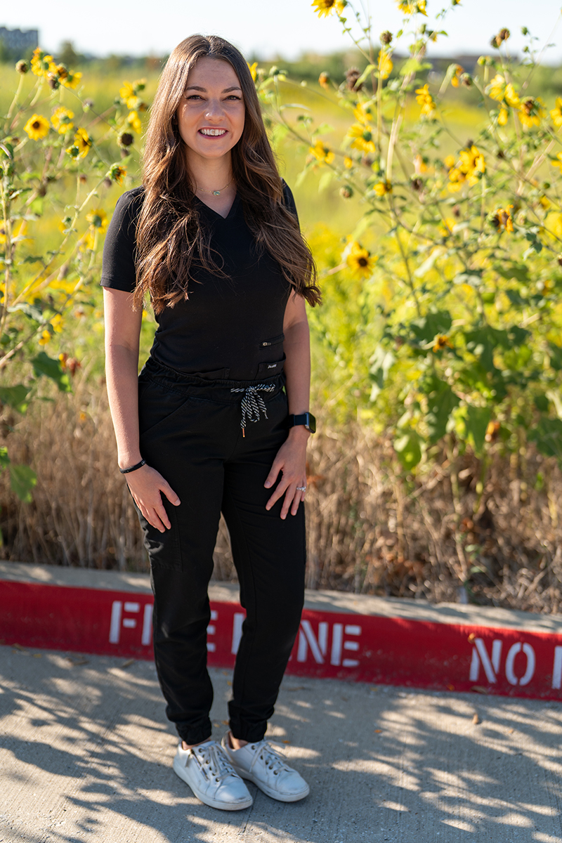 A woman with long brown hair, wearing black scrubs and white sneakers, stands and smiles outdoors in front of sunflower plants.