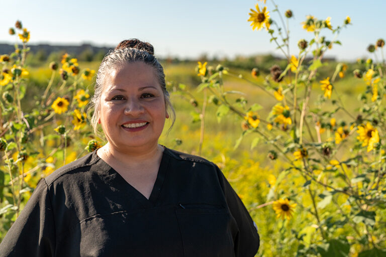 A woman in black scrubs stands outdoors in front of tall sunflowers, smiling at the camera on a sunny day.