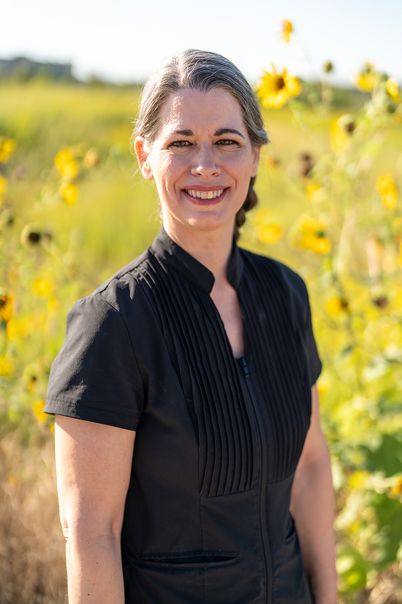 A woman with gray-streaked hair wearing a black short-sleeved top stands outdoors in front of tall green grass and yellow flowers, smiling at the camera.