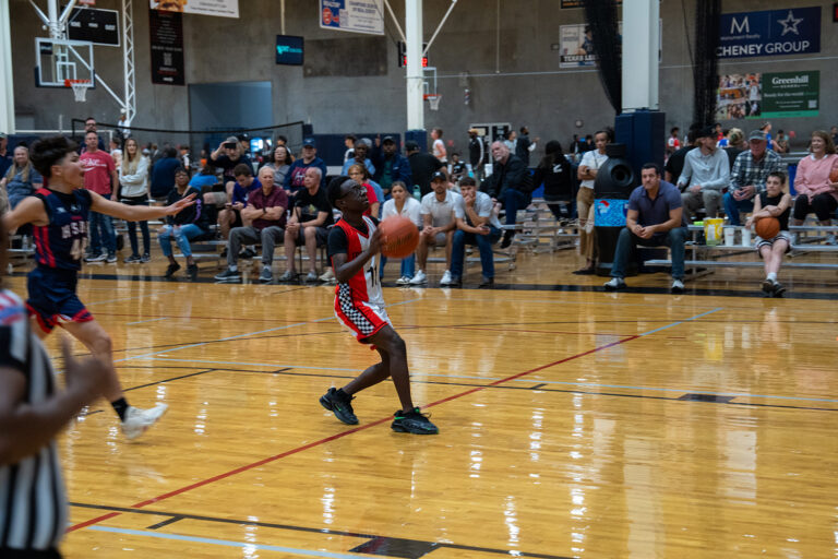 A youth basketball player in a red and black uniform dribbles on an indoor court as opponents and spectators watch from the bleachers.