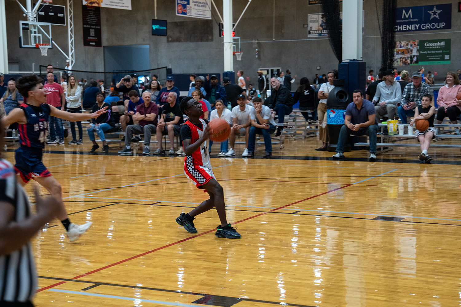A youth basketball player in a red and black uniform dribbles on an indoor court as opponents and spectators watch from the bleachers.