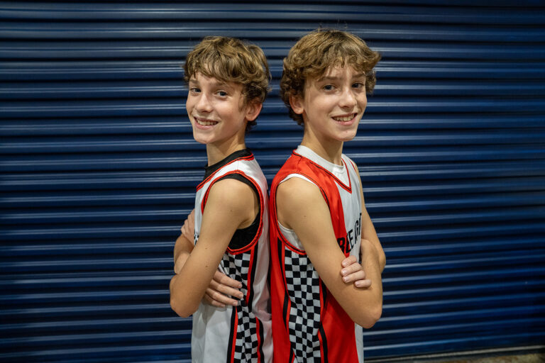 Two boys with similar appearances stand back to back, arms crossed, wearing basketball jerseys with checkered side panels in front of a blue metal shutter.
