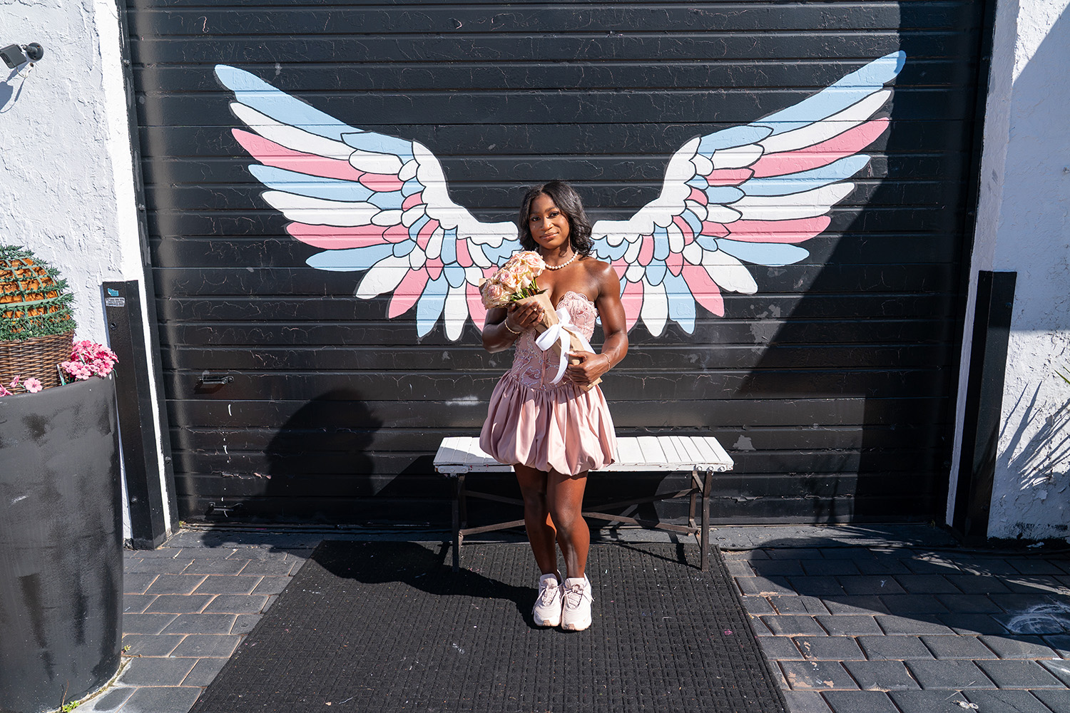 A woman in a pink dress holding flowers stands in front of a mural of large angel wings painted on a black wall.